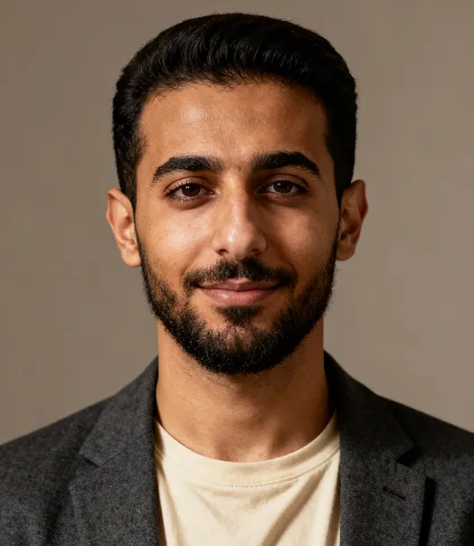 Portrait of a young man with dark hair and beard, wearing a beige shirt and gray blazer, smiling slightly against a plain beige background.
