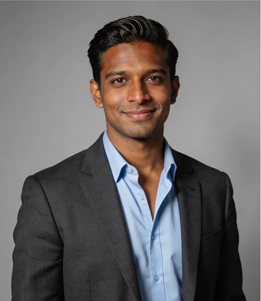Portrait of a confident young man wearing a light blue shirt and dark blazer against a gray background.