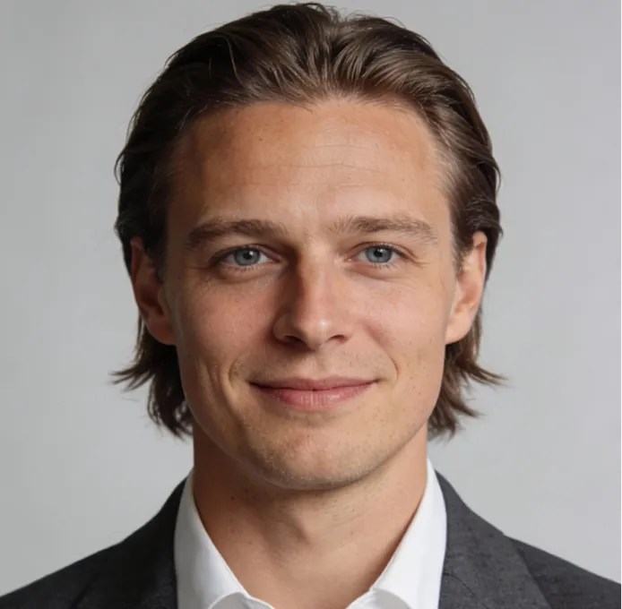 Portrait of a young man with light brown hair, blue eyes, wearing a white shirt and gray blazer, smiling slightly against a gray background.
