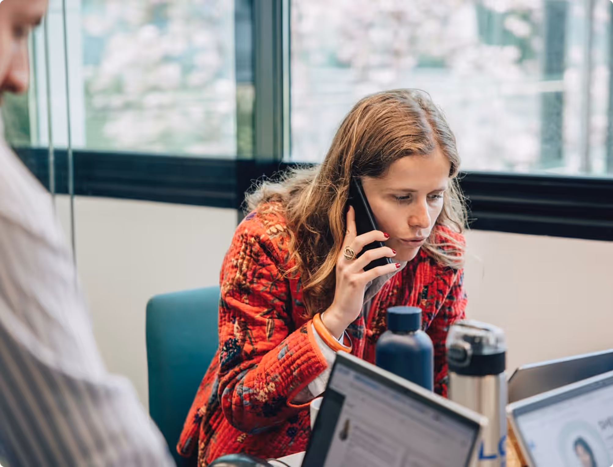 Femme en veste rouge parlant au téléphone dans un bureau avec un ordinateur portable devant elle.