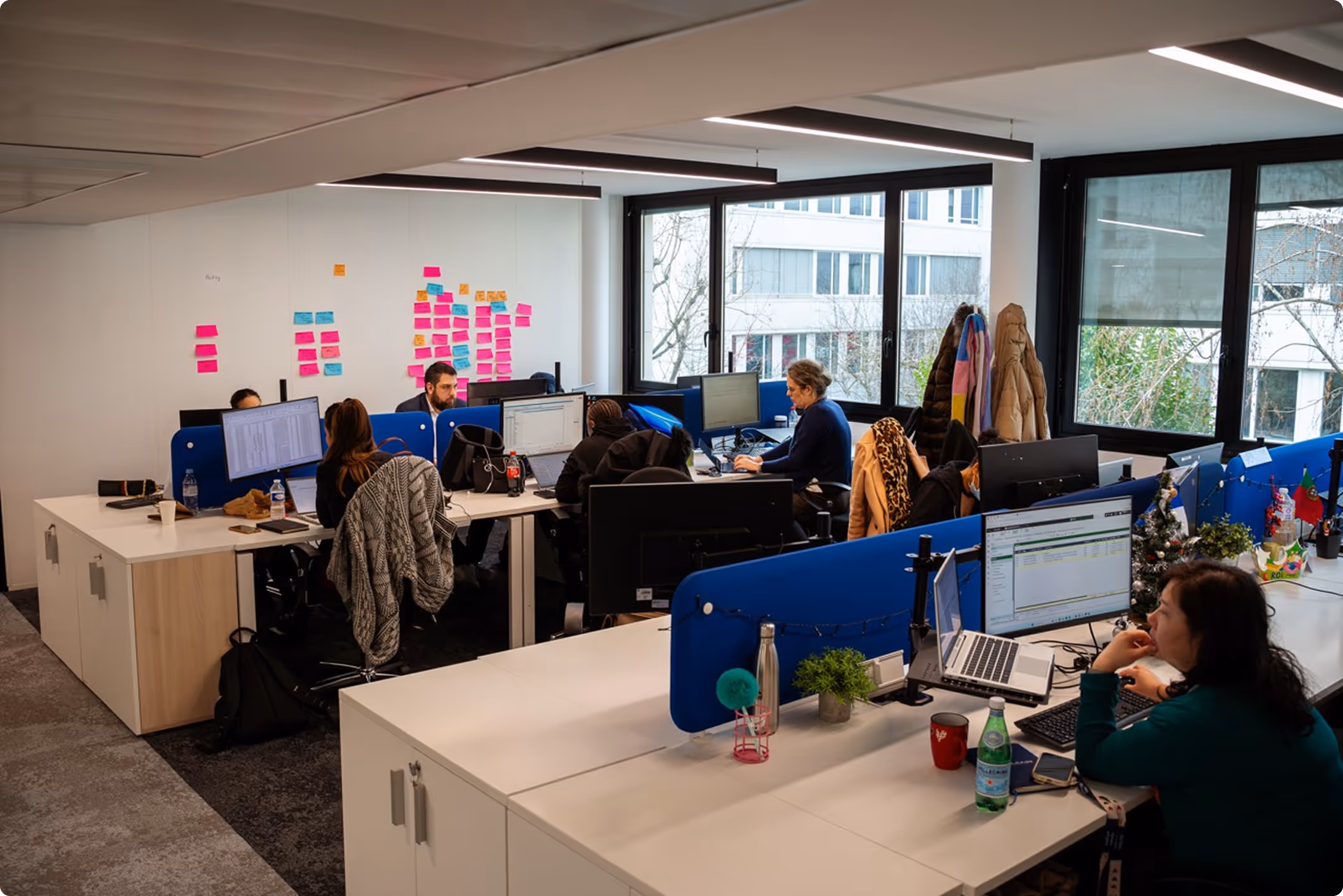 Bureau moderne avec plusieurs employés travaillant sur des ordinateurs, murs avec notes autocollantes colorées, grandes fenêtres laissant entrer la lumière naturelle.