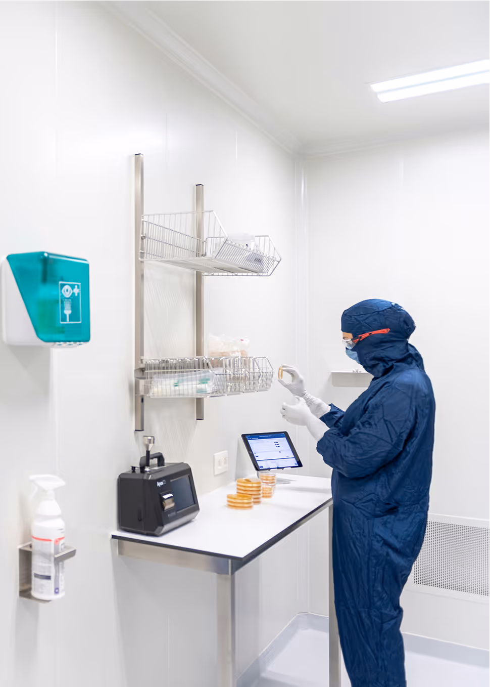 Technicien en blouse bleue inspectant une boîte de Pétri dans un laboratoire stérile avec équipement de laboratoire et écran tactile.