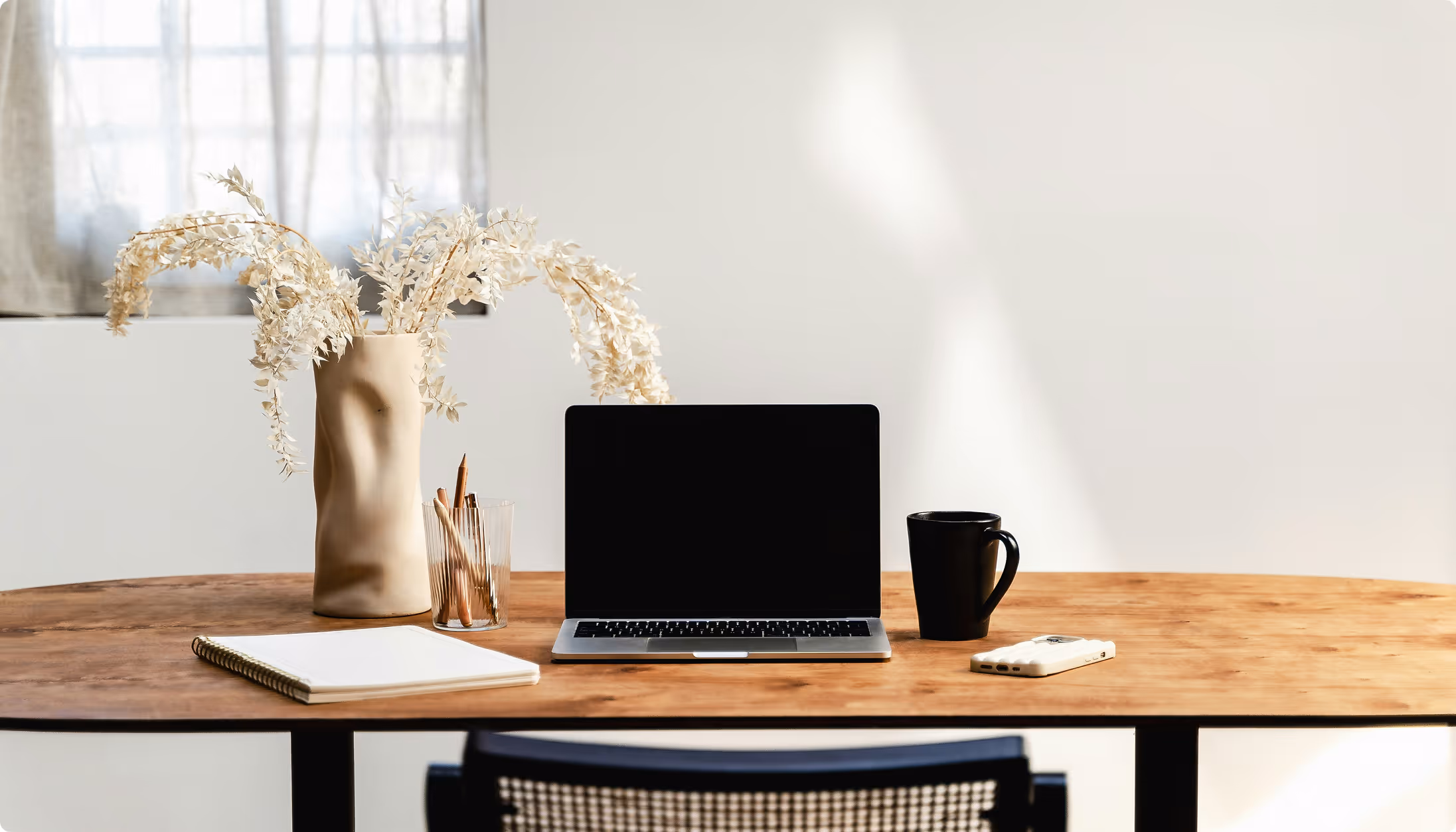 Bureau en bois avec un ordinateur portable fermé, un carnet, un pot avec des crayons, une tasse noire et un vase beige avec des branches séchées.