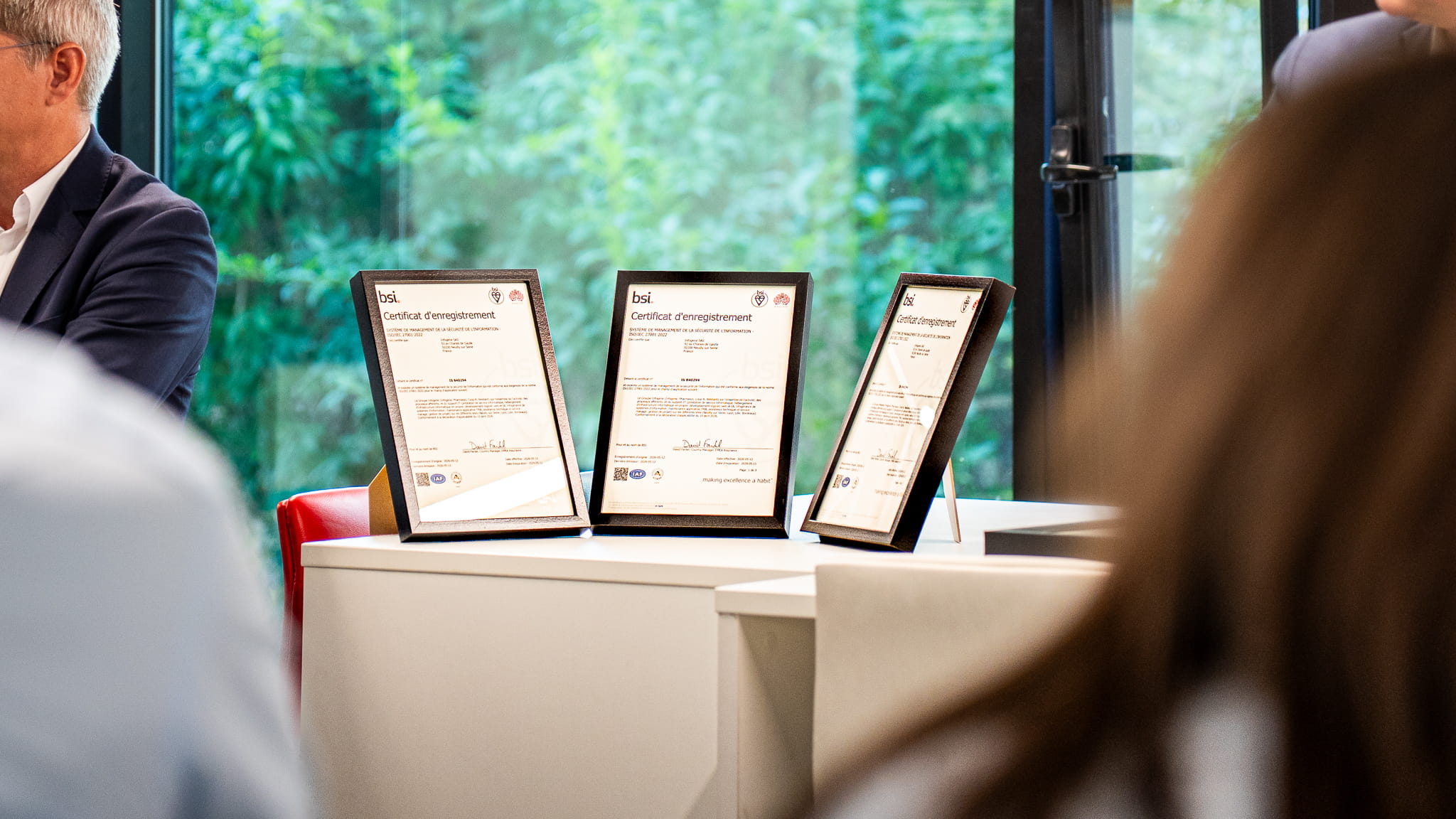Trois certificats d'enregistrement BSI encadrés posés sur un bureau blanc près d'une fenêtre.