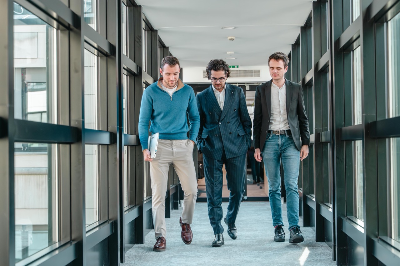 Trois hommes marchant dans un couloir vitré, discutant en milieu professionnel.
