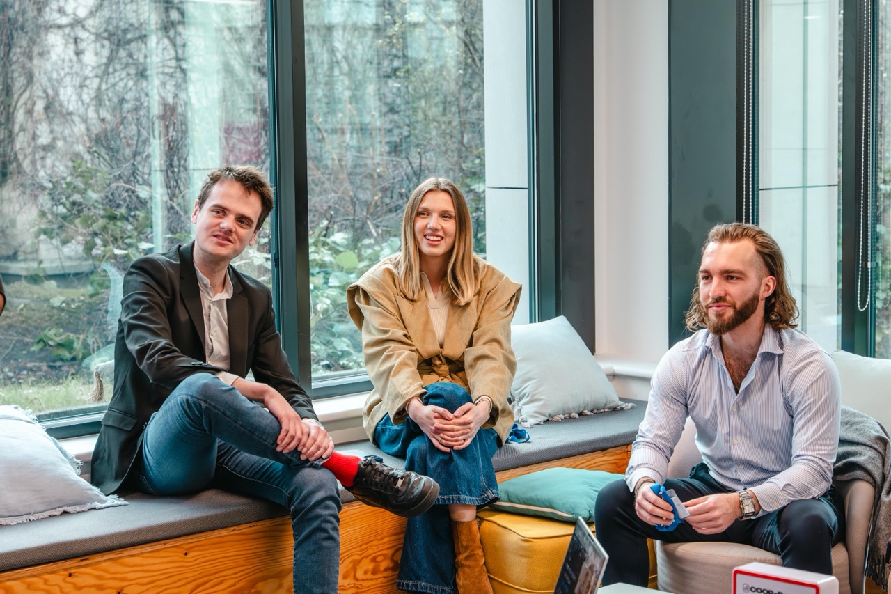 Trois jeunes professionnels assis, souriants, en discussion dans un espace de travail lumineux avec grandes fenêtres.