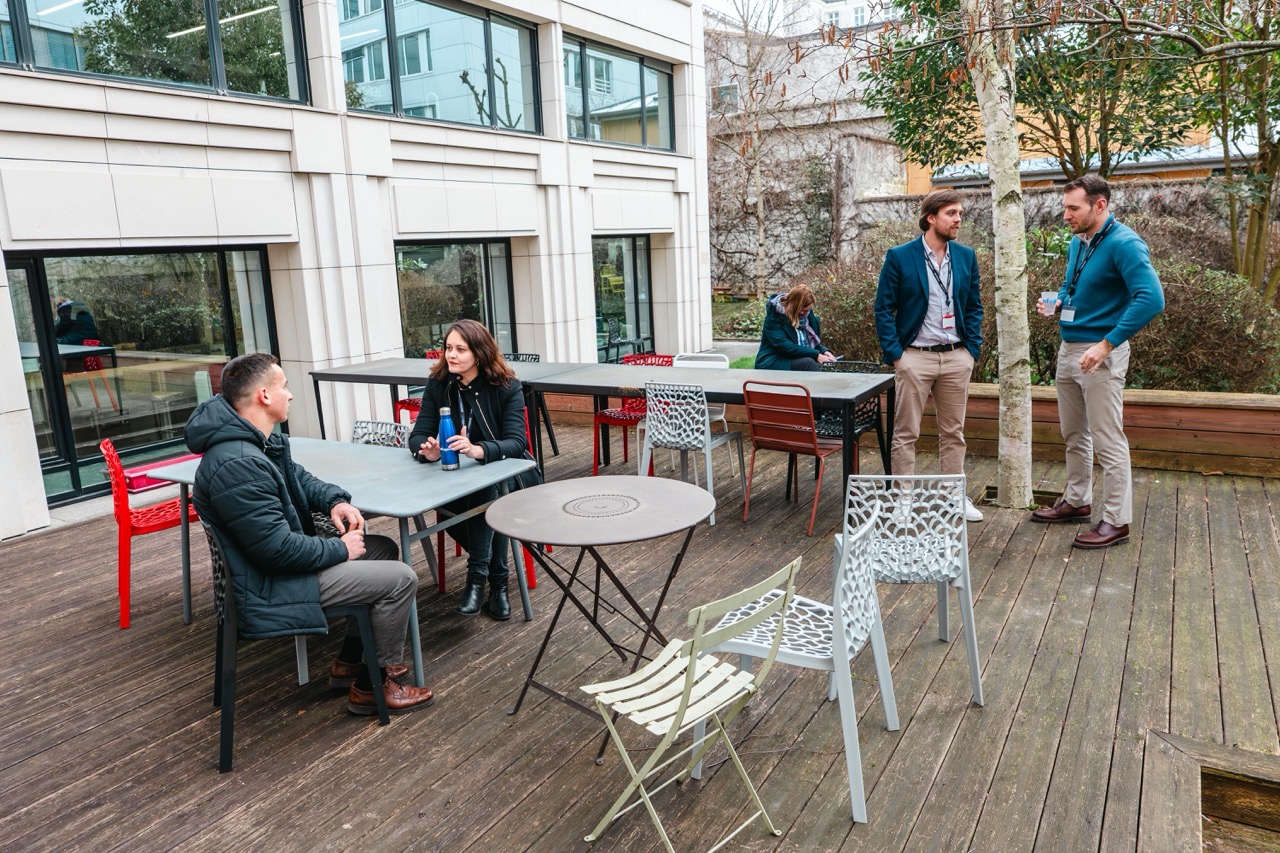 Personnes discutant assises et debout sur une terrasse en bois avec des tables et des chaises.