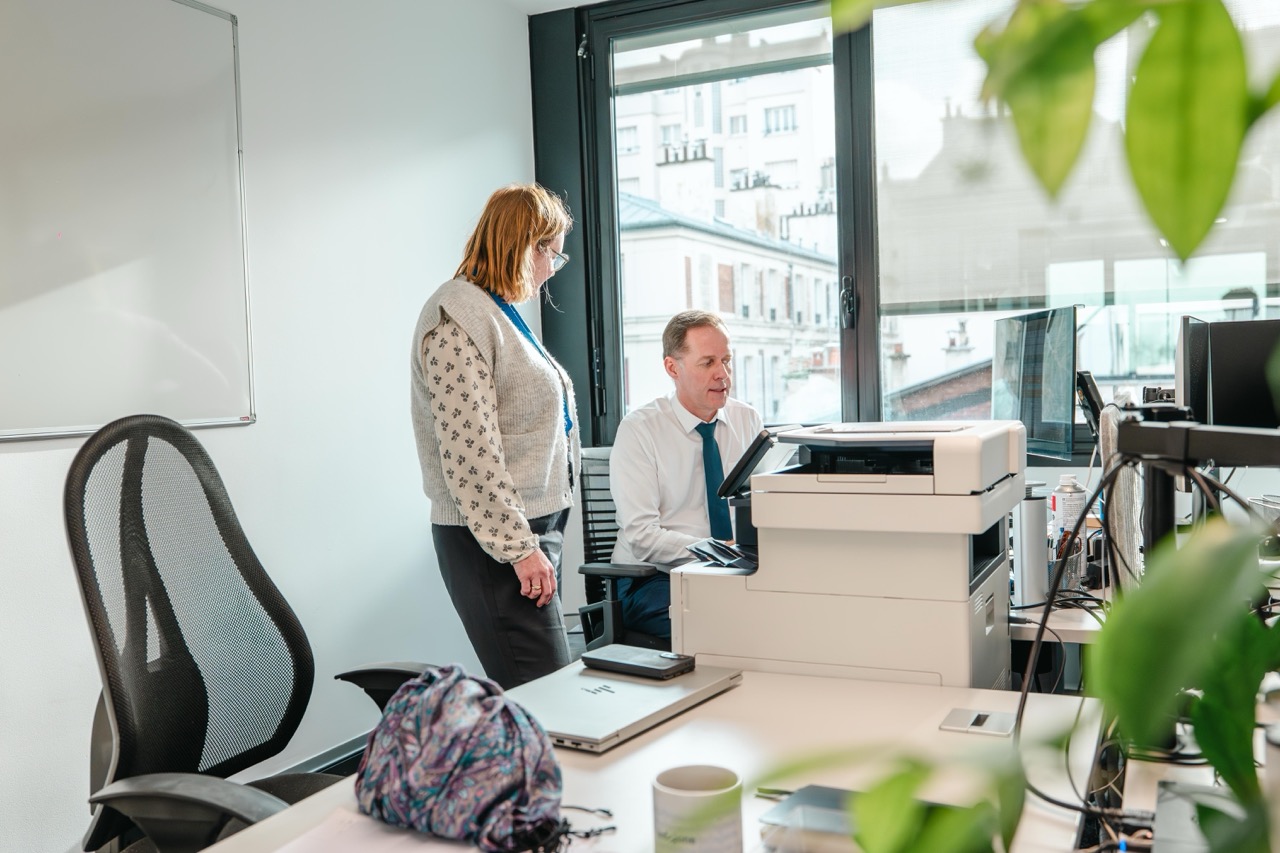Un homme assis et une femme debout discutent près d'une imprimante dans un bureau lumineux.