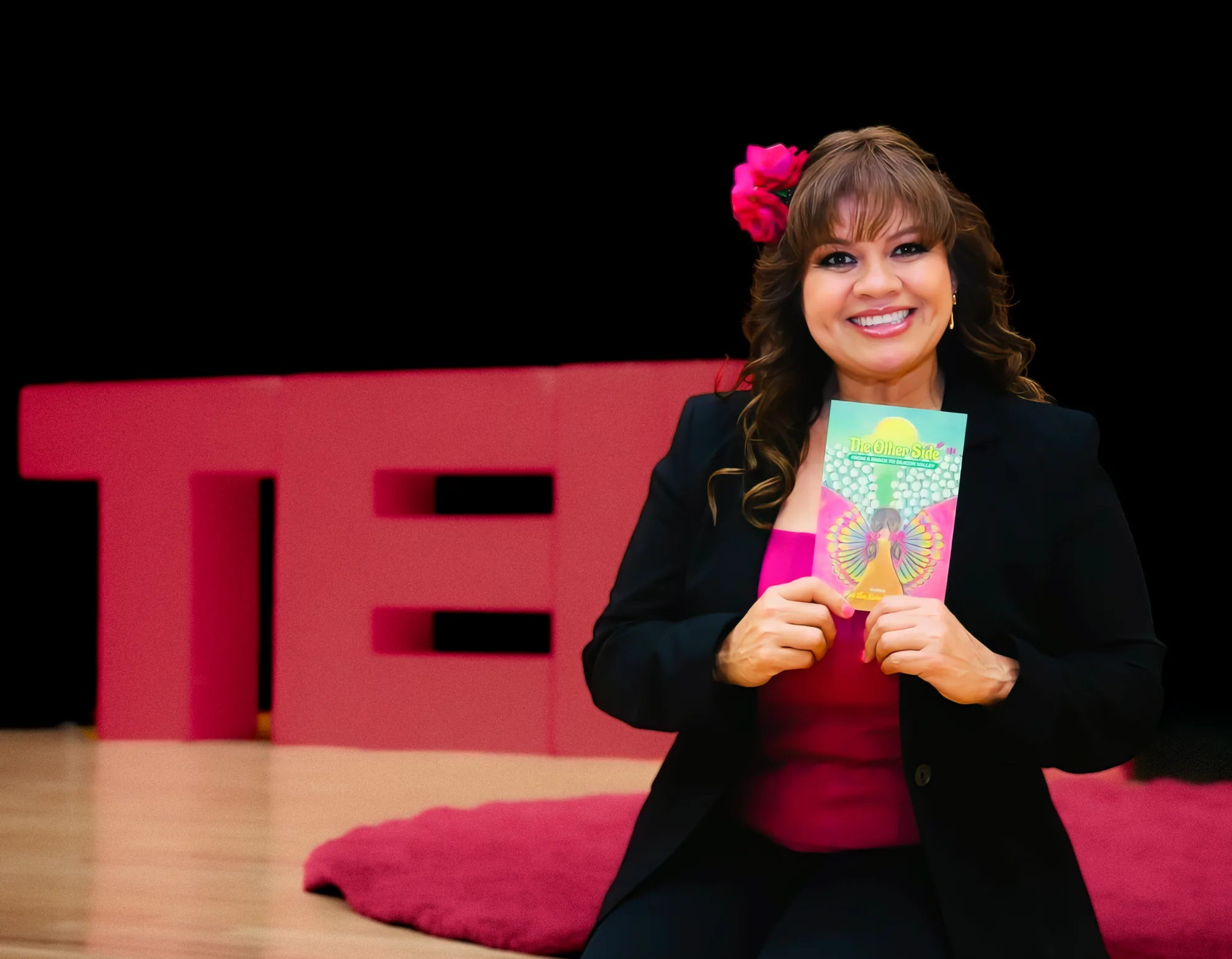 Martha Niño Rodriguez seated on a TEDx stage, holding her book The Other Side