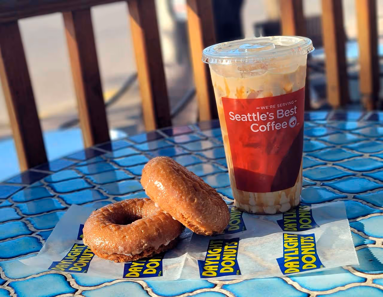 Two glazed donuts on a Daylight Donuts wrapper next to a plastic cup of iced Seattle's Best Coffee on a blue tiled table.