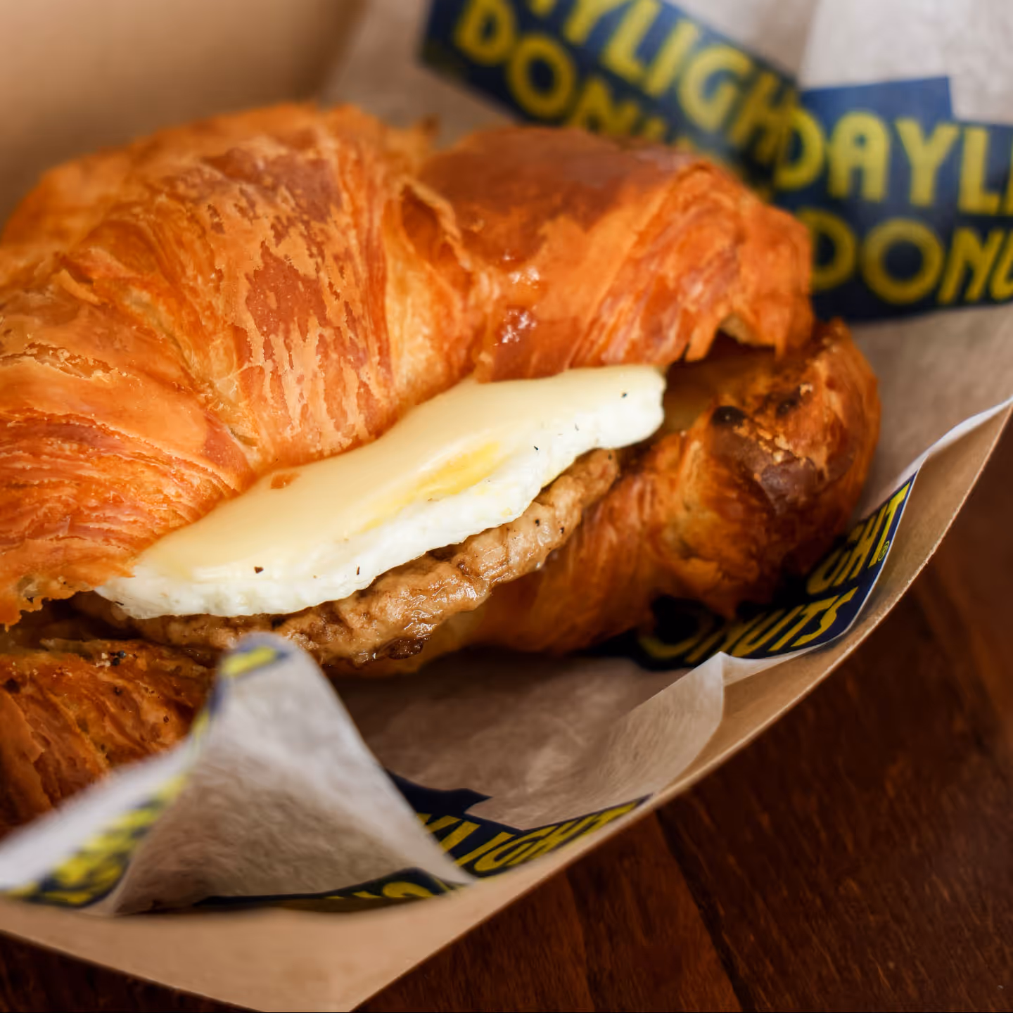 Croissant sandwich with fried egg and sausage patty on branded parchment paper.