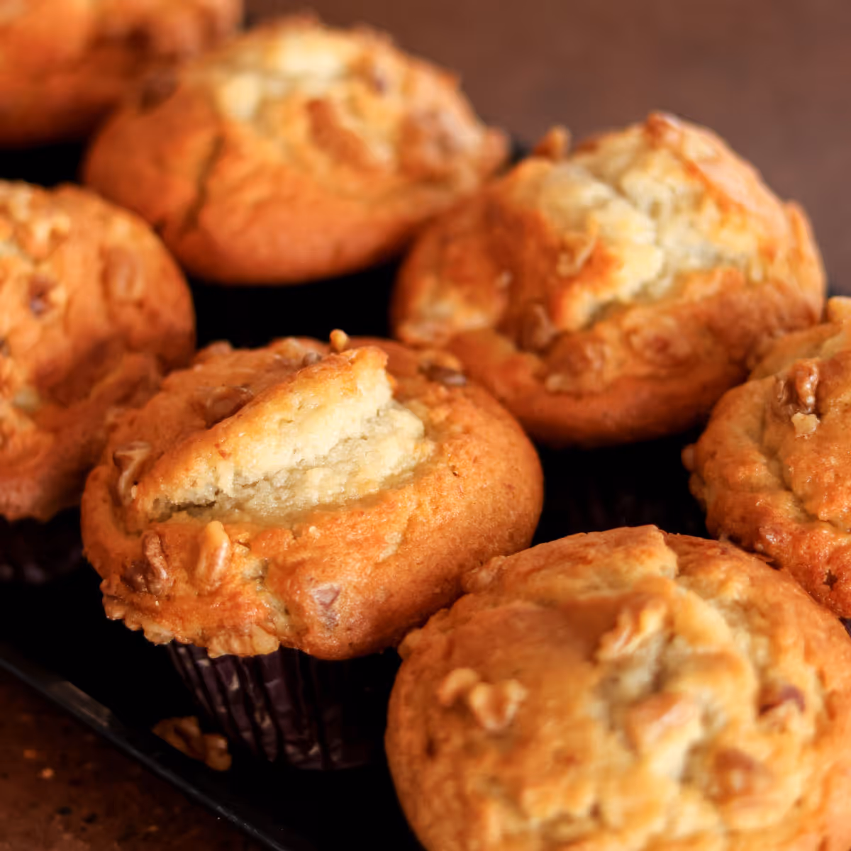 Close-up of several golden-brown muffins with a cracked top and walnuts on a dark surface.