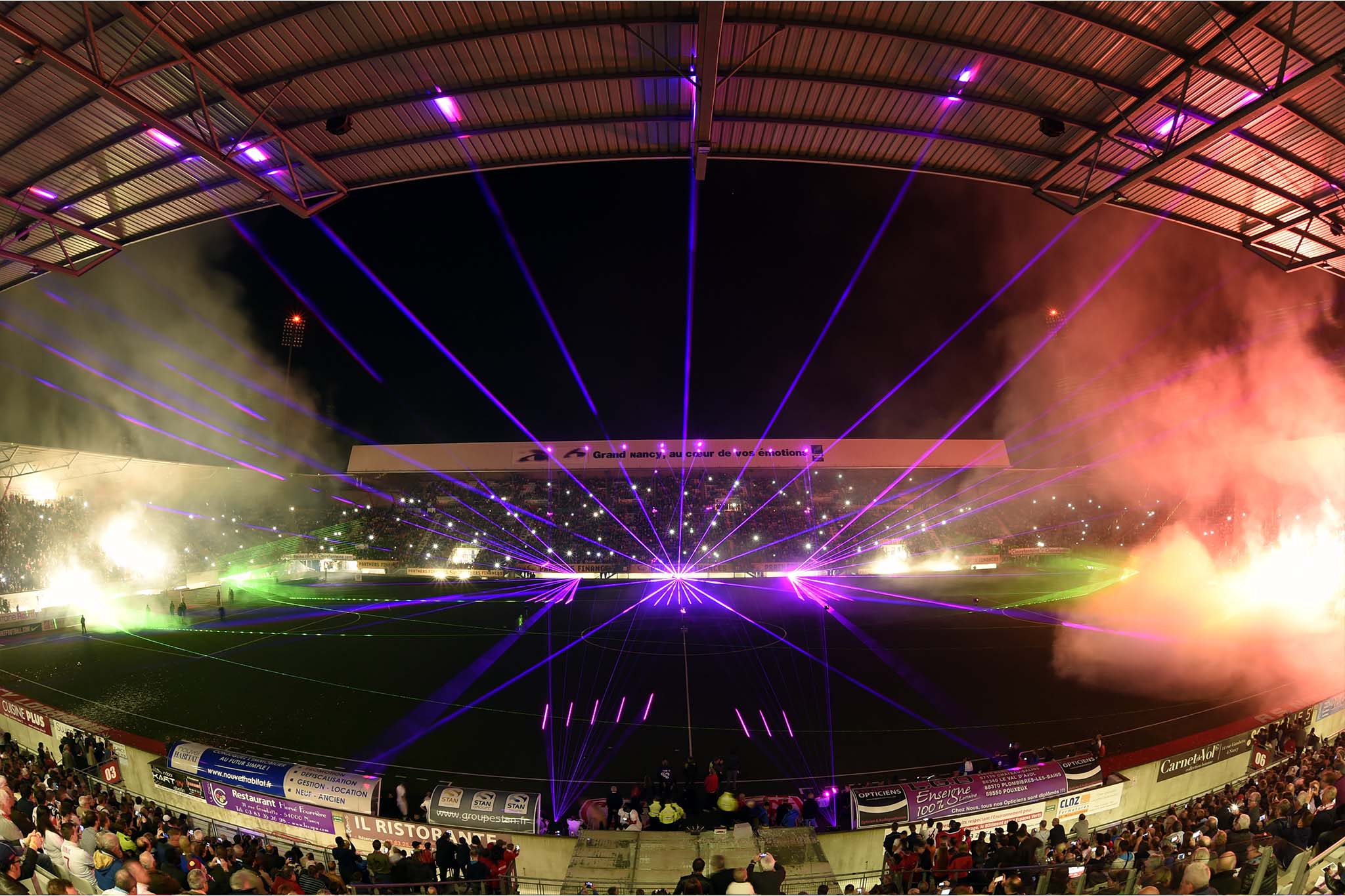 Stade de football nocturne illuminé par des effets laser colorés et fumée sur le terrain devant des milliers de spectateurs