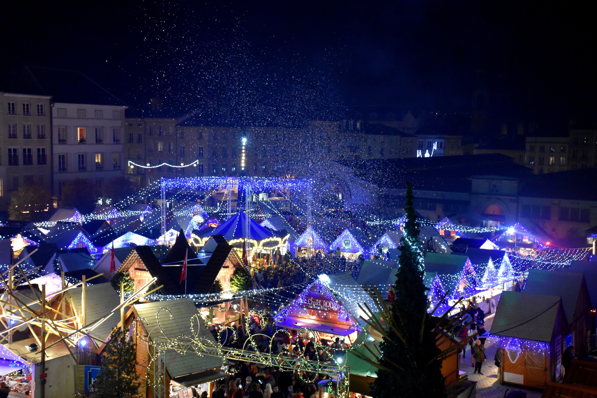 Marché de Noël de Nancy la nuit avec stands illuminés lumières festives et neige artificielle tombant sur la foule