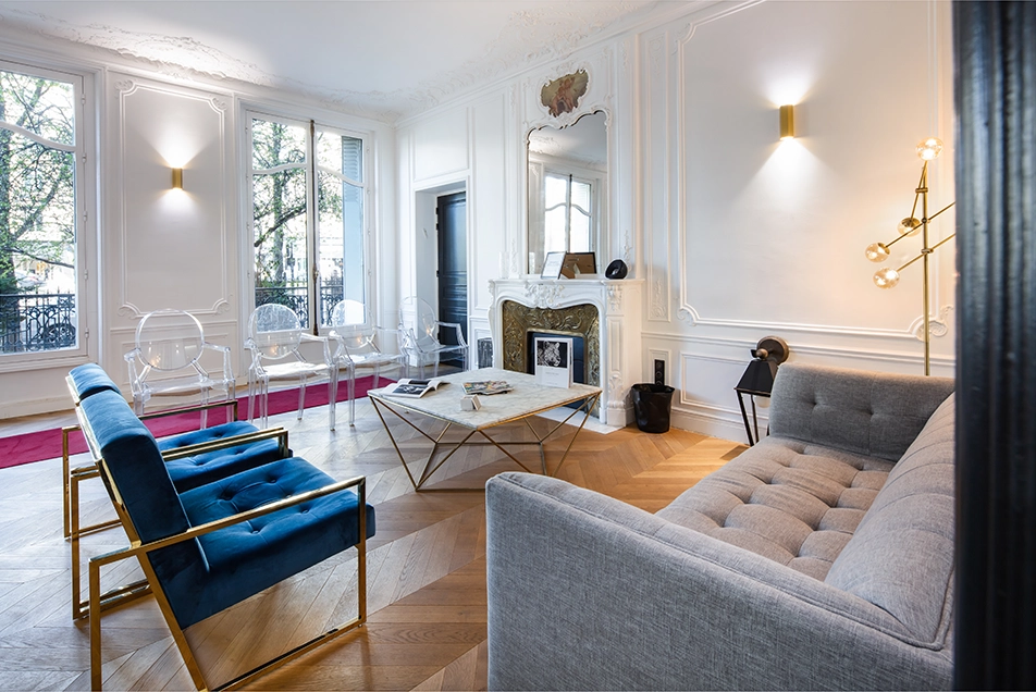 Salon élégant avec fauteuils bleus, chaises transparentes, canapé gris et cheminée ornée.