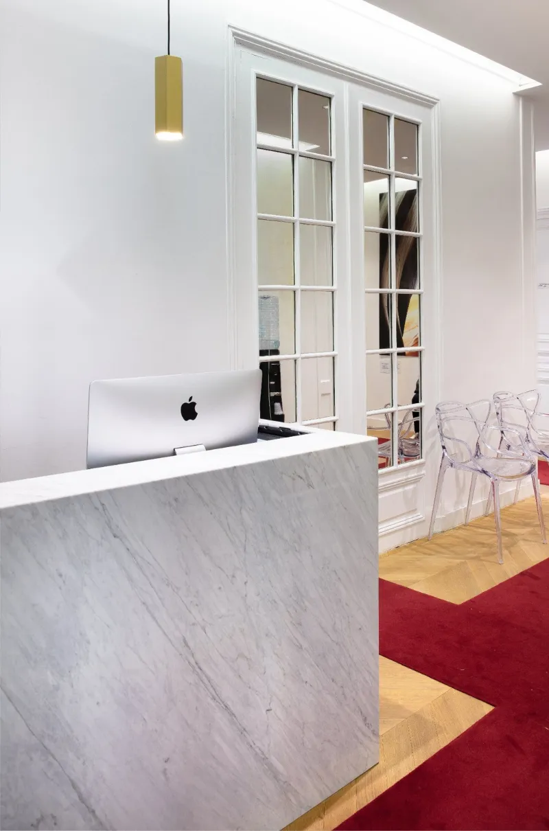 Réception avec bureau en marbre, ordinateur Apple, chaises transparentes et miroir encadré au mur.
