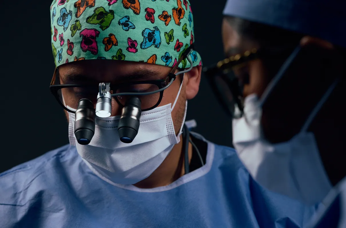 Le chirurgien Adrien Fellous portant un masque, une coiffe colorée et des lunettes avec loupe pendant une opération.