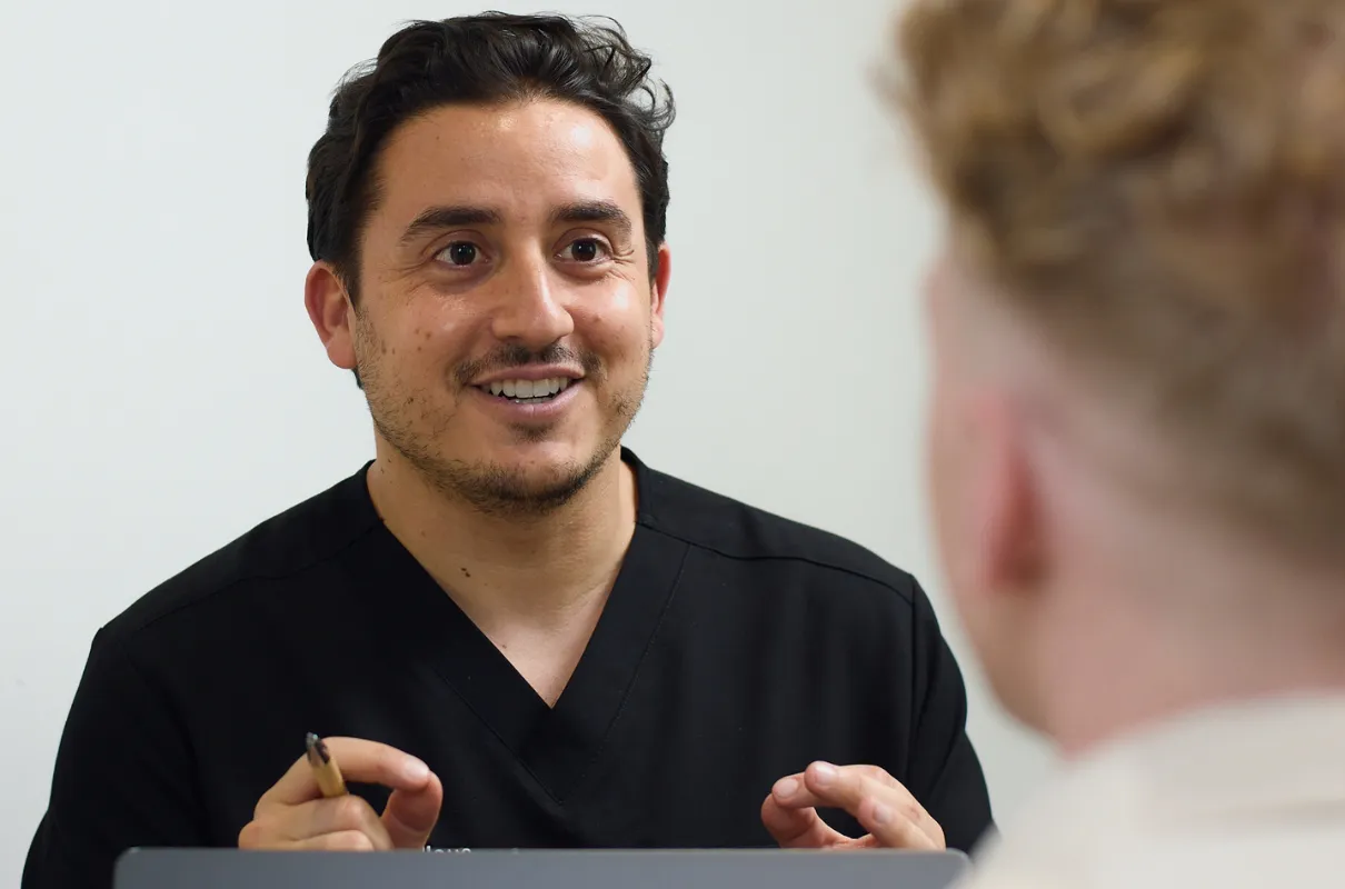 Dr Adrien Fellous en blouse noire souriant et discutant avec une autre personne floue au premier plan.