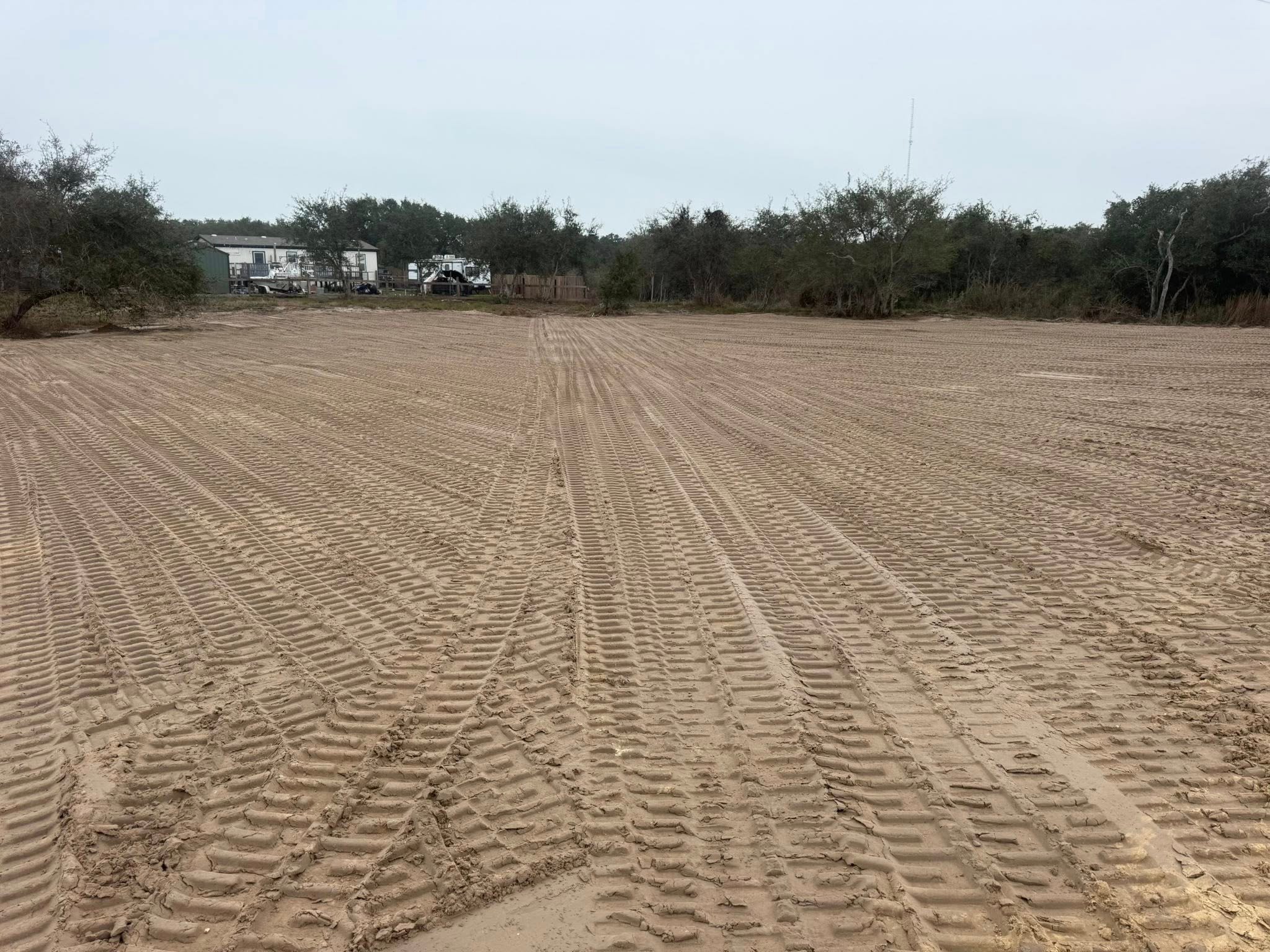 Large cleared and leveled dirt field with visible tire tracks, bordered by trees and some buildings in the distance under an overcast sky.