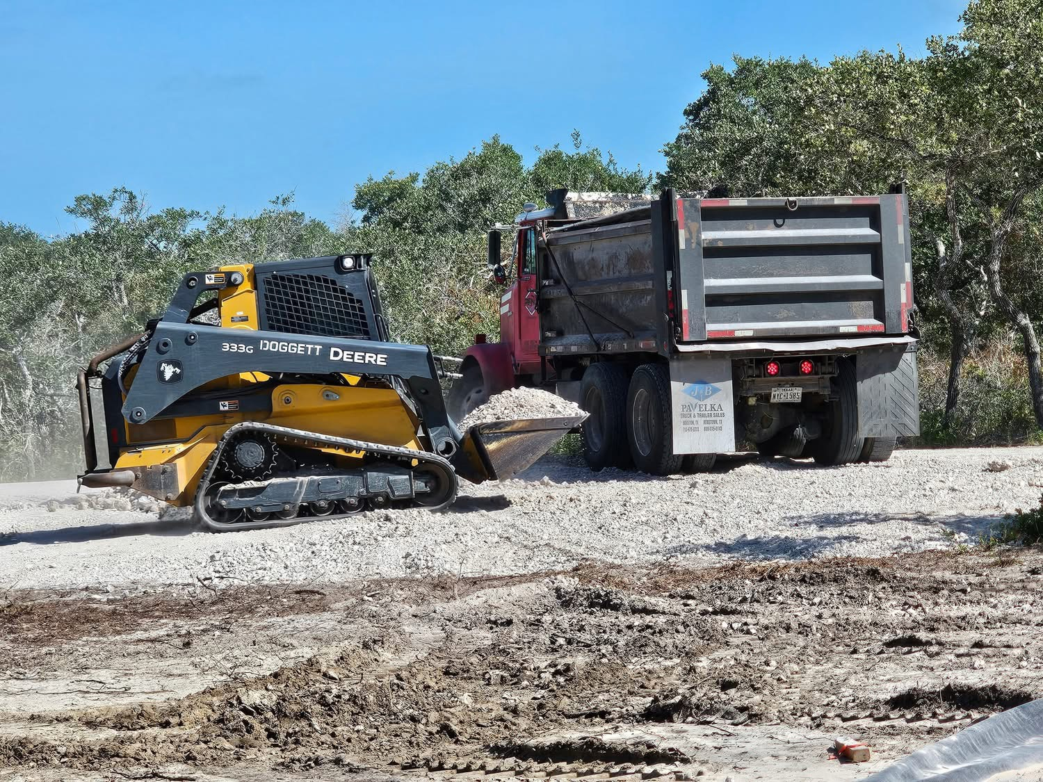 Yellow John Deere 333G skid steer loader dumping gravel into the back of a red dump truck on a construction site with trees in the background.