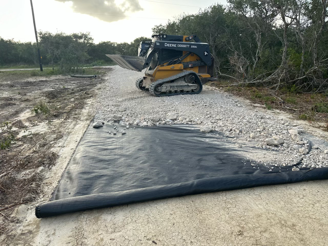 Yellow John Deere 333G compact track loader on a gravel-covered construction path near a black geotextile fabric.