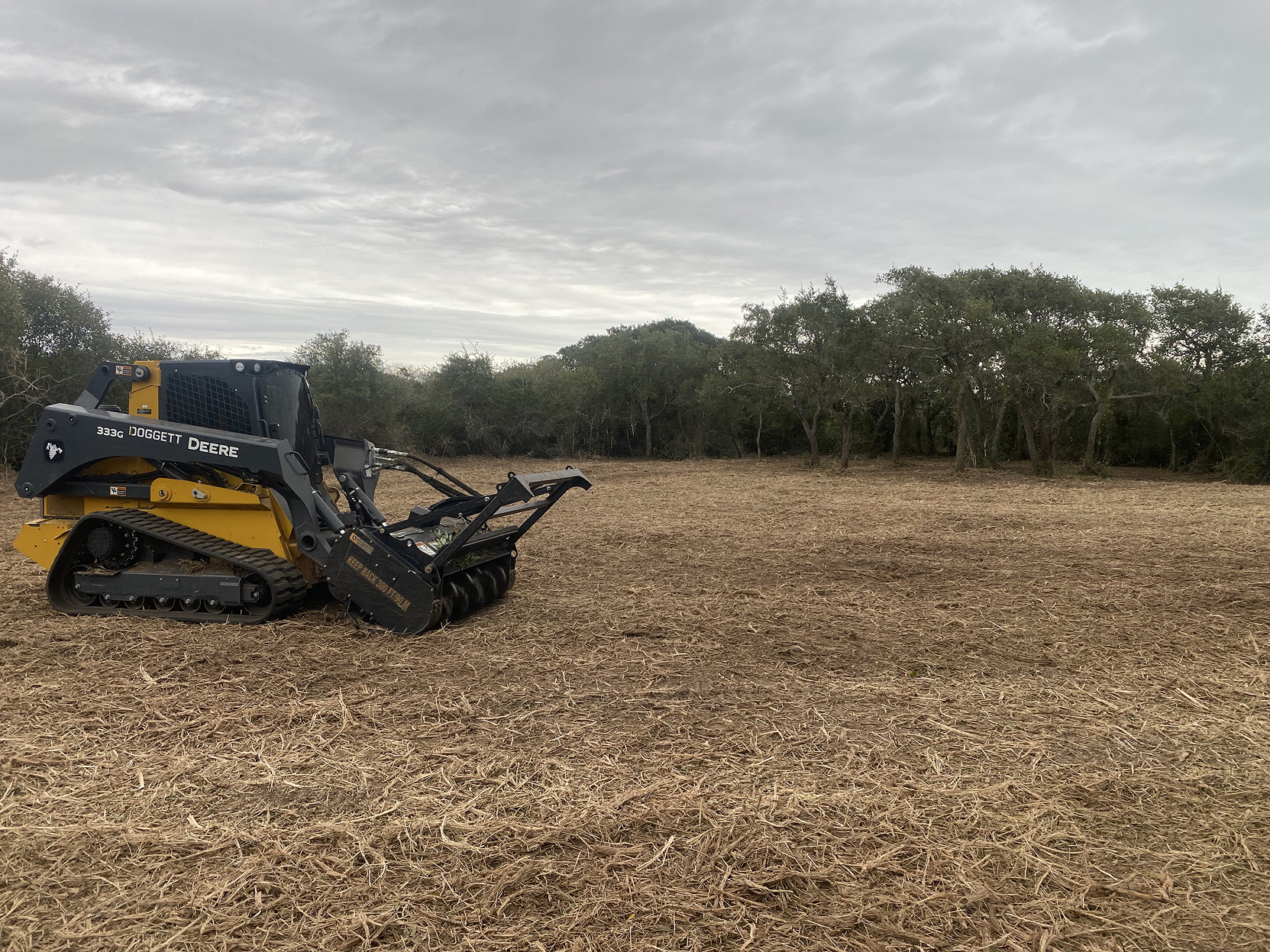 Yellow and black John Deere 333G skid steer with soil tiller attachment on a cleared field under a cloudy sky with trees in the background.
