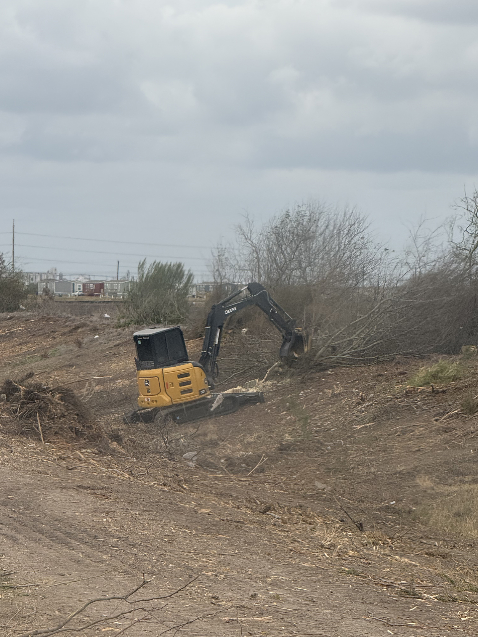 Yellow John Deere excavator clearing brush and small trees on a dirt construction site under a cloudy sky.