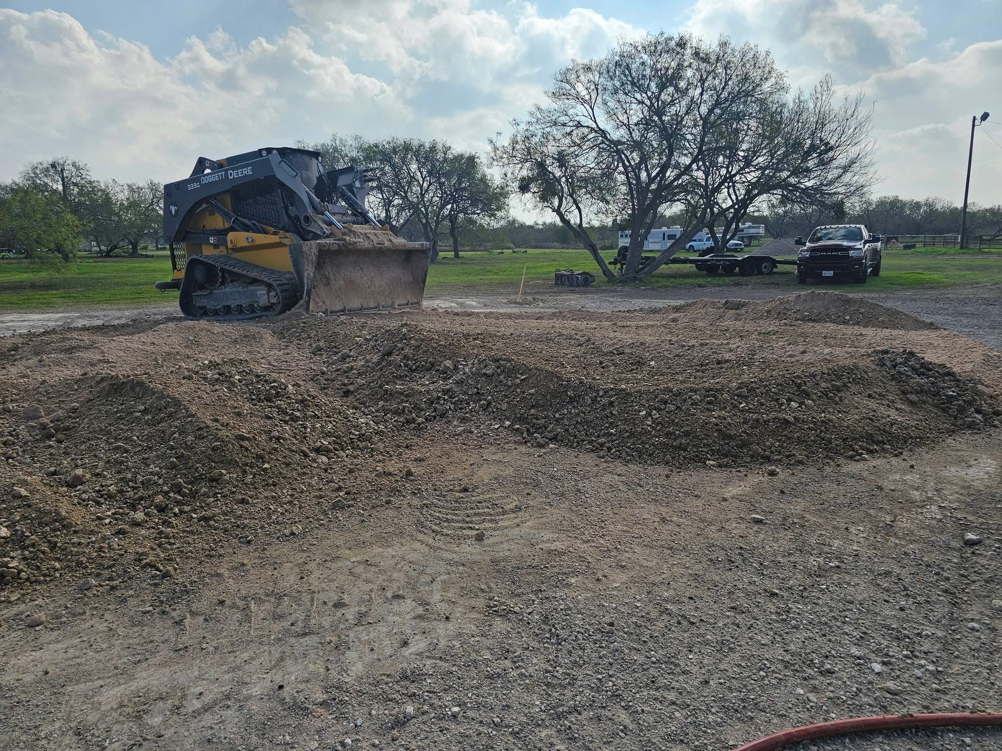 John Deere 333G compact track loader moving dirt at a construction site with trees and vehicles in the background under a partly cloudy sky.