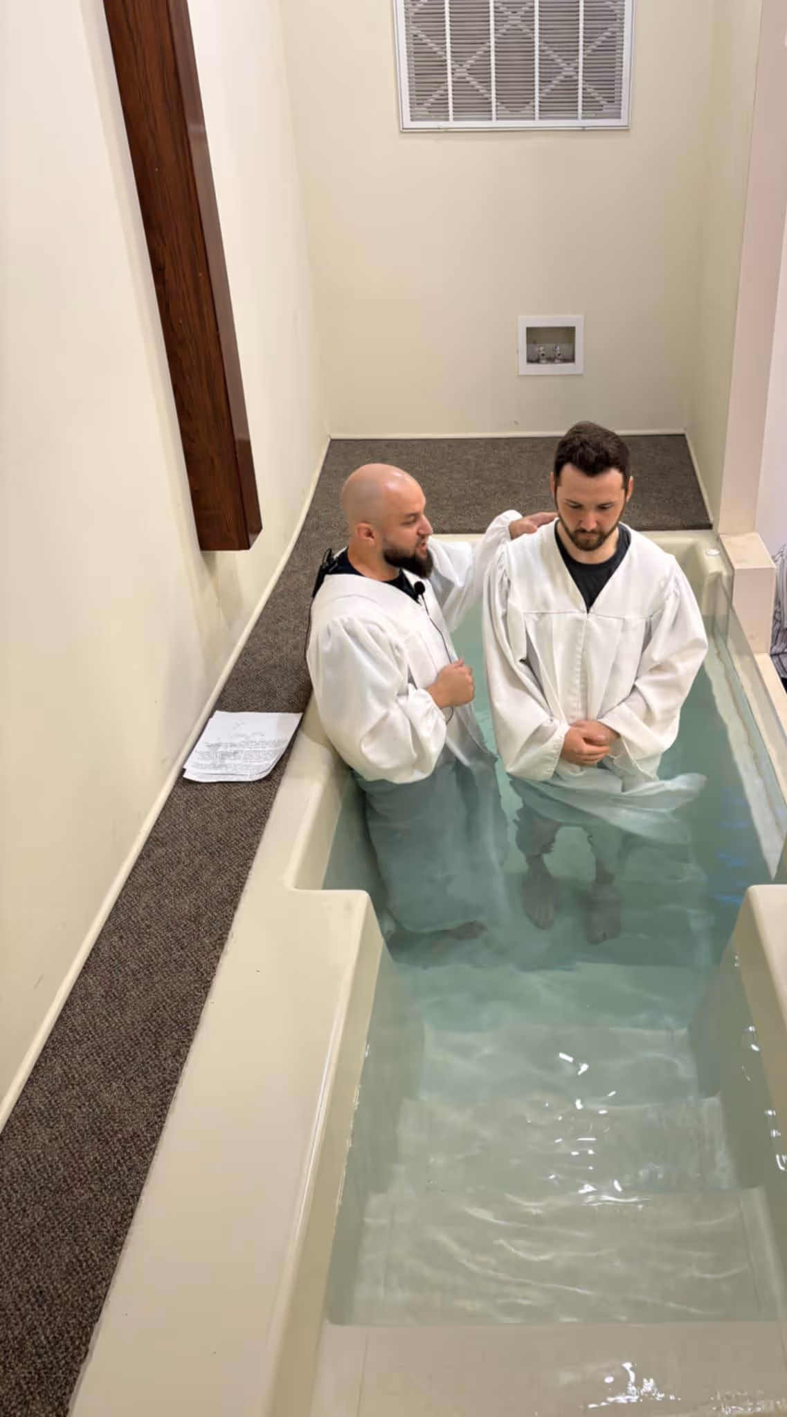 Two men wearing white robes standing in a baptismal pool, one with hand on the other's shoulder.
