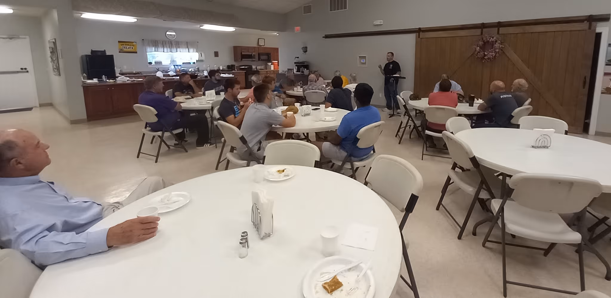 Group of people seated at round tables in a room, listening to a person speaking near a wooden wall.