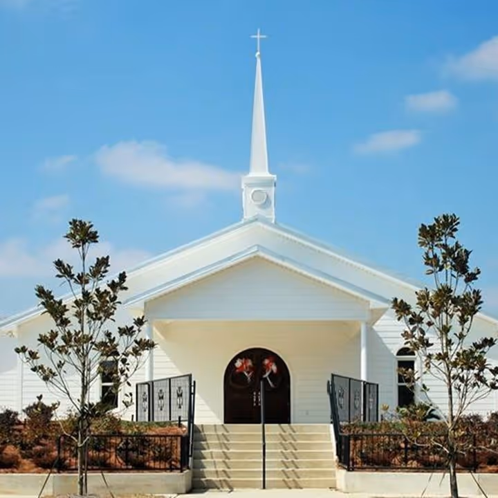 White church with a central steeple, cross, stairs, and two small trees against a blue sky.