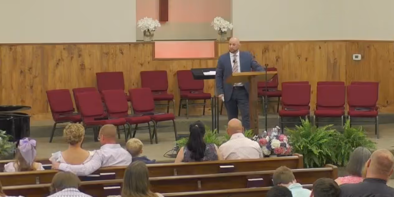 Man in suit speaking at a wooden lectern in a church with seated audience and red chairs behind him.