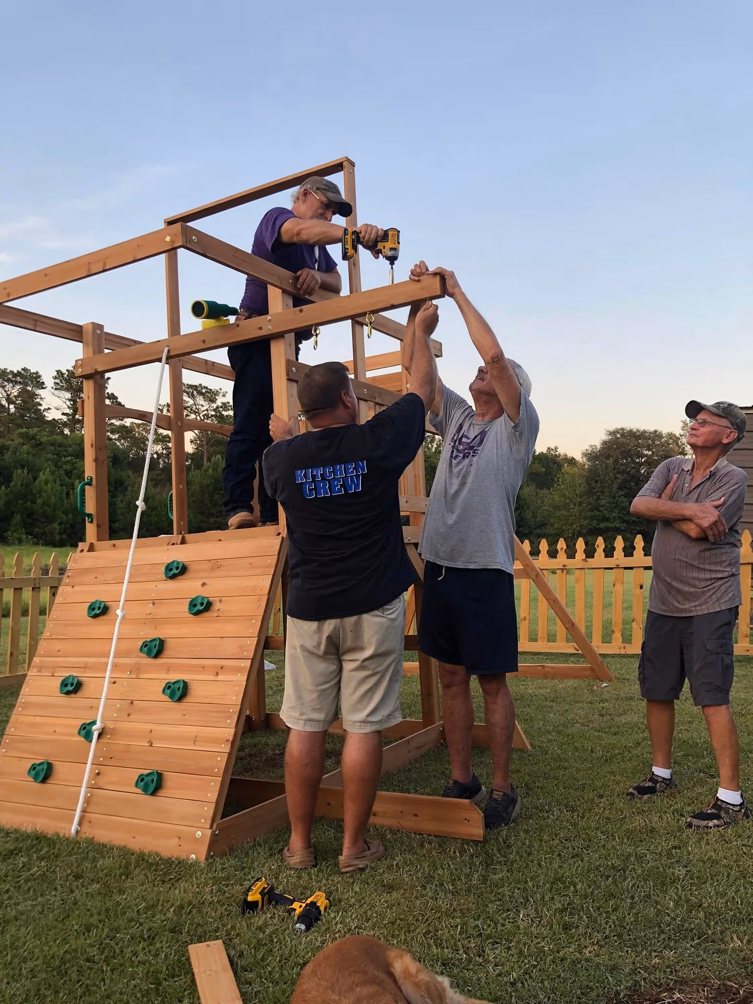 Four men assembling a wooden play structure with climbing wall outdoors in a fenced yard.