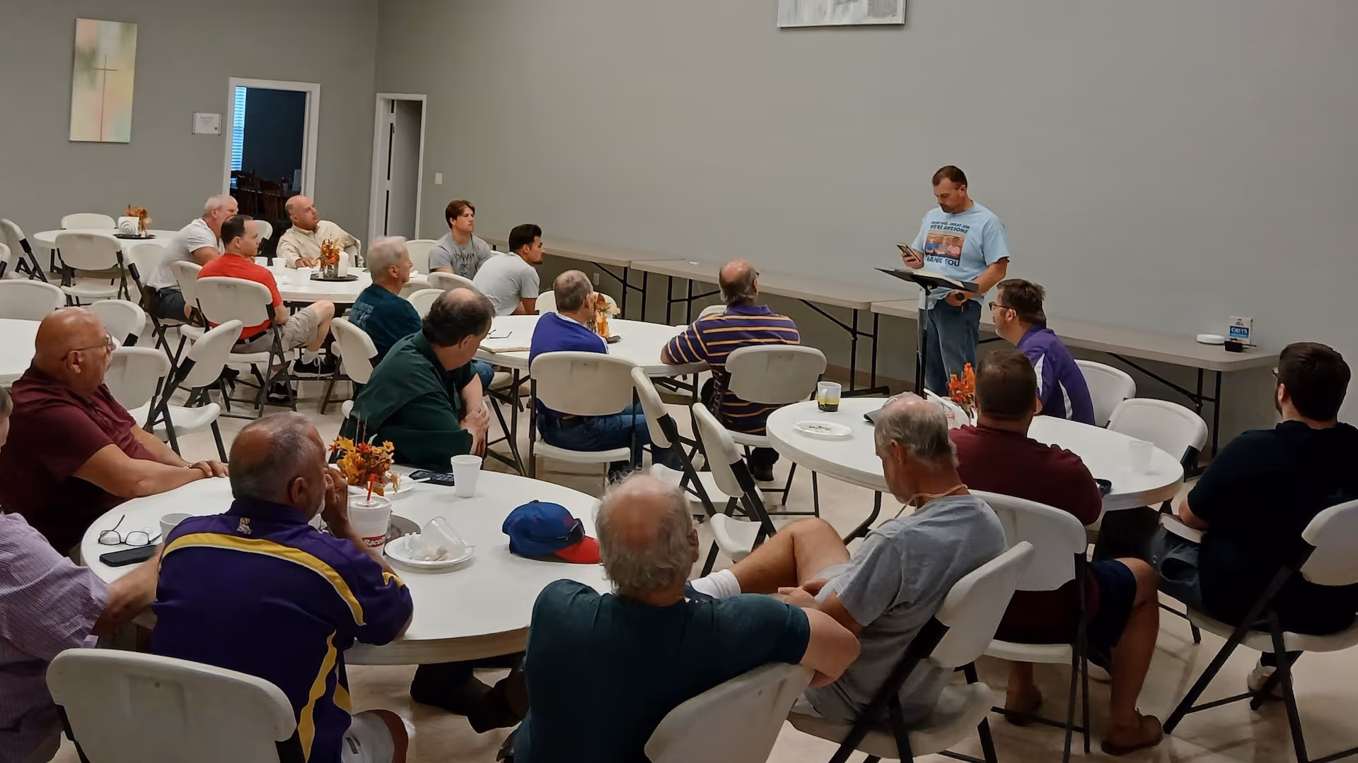 Group of men seated around tables listening to a man standing and speaking in a room.