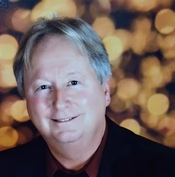 Smiling older man with gray hair wearing a dark suit and burgundy shirt, with a bokeh background.