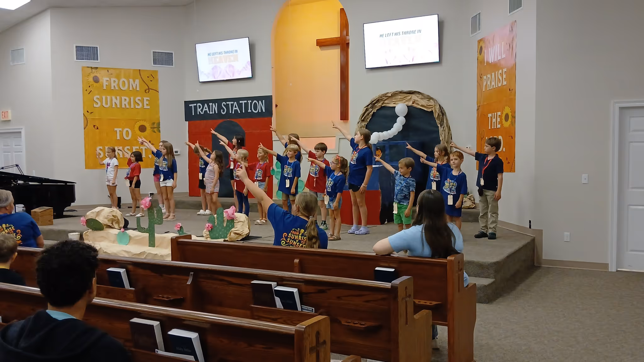 Group of children on stage in church pointing upwards during a performance with themed decorations.