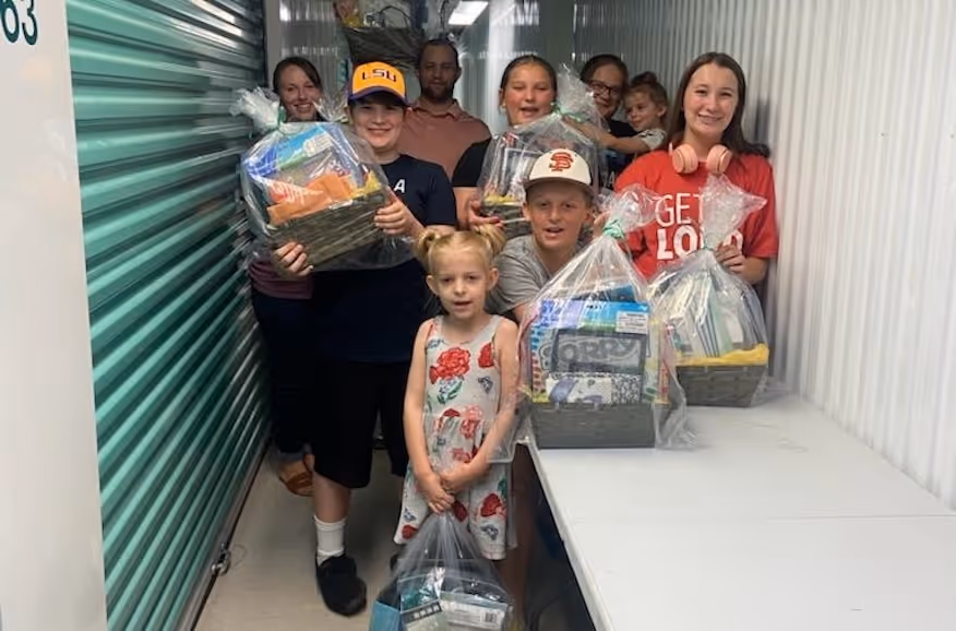 Group of smiling children and adults holding gift baskets wrapped in plastic in a hallway with storage units.