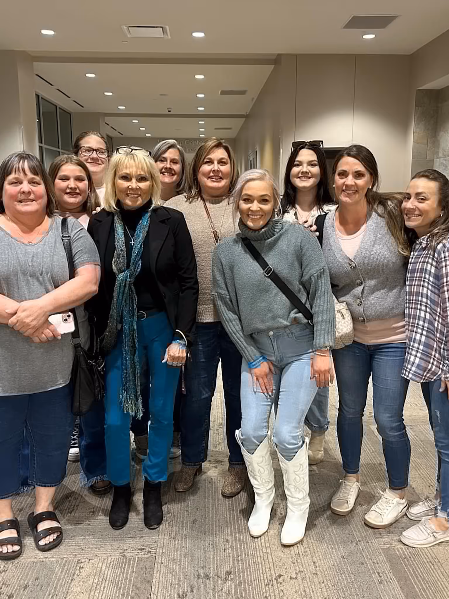Group of ten smiling women standing close together indoors, dressed casually in sweaters and jeans.