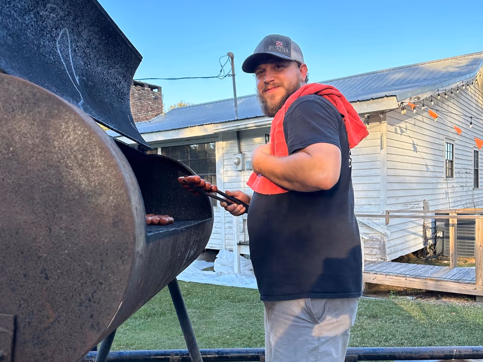 Man in black shirt and gray hat grilling sausages outdoors near a white house with a red towel on shoulder.