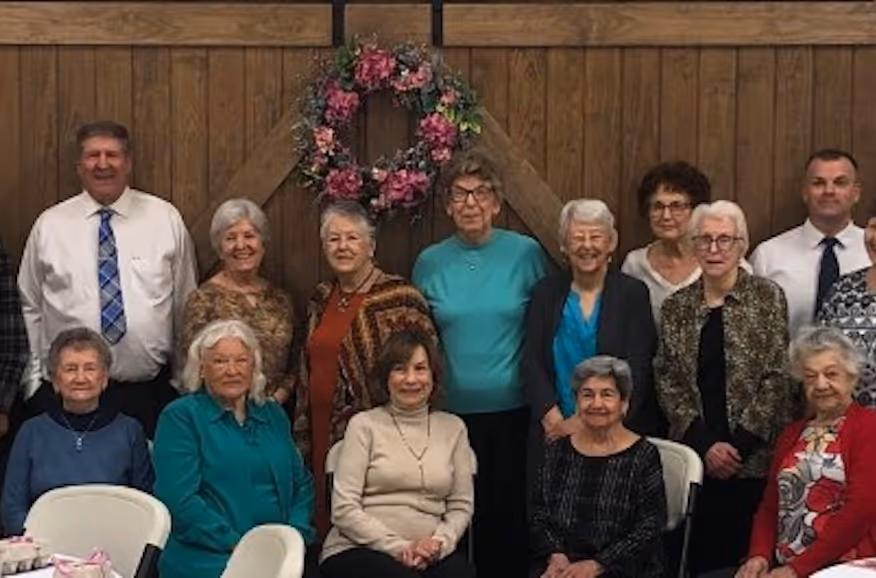 Group of elderly people posing indoors with a floral wreath on wood wall behind them.
