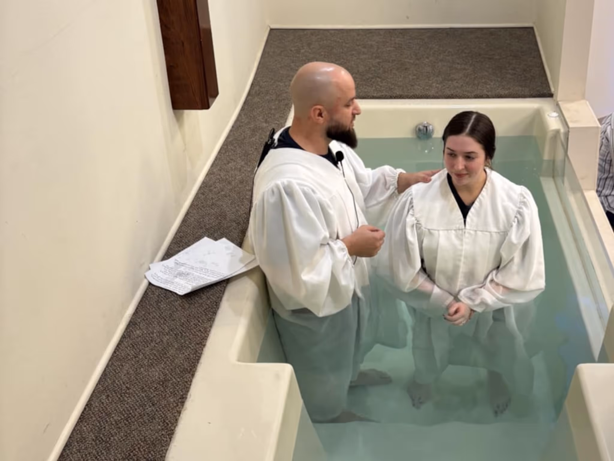 Man and woman in white robes standing in baptismal pool inside church during baptism ceremony.