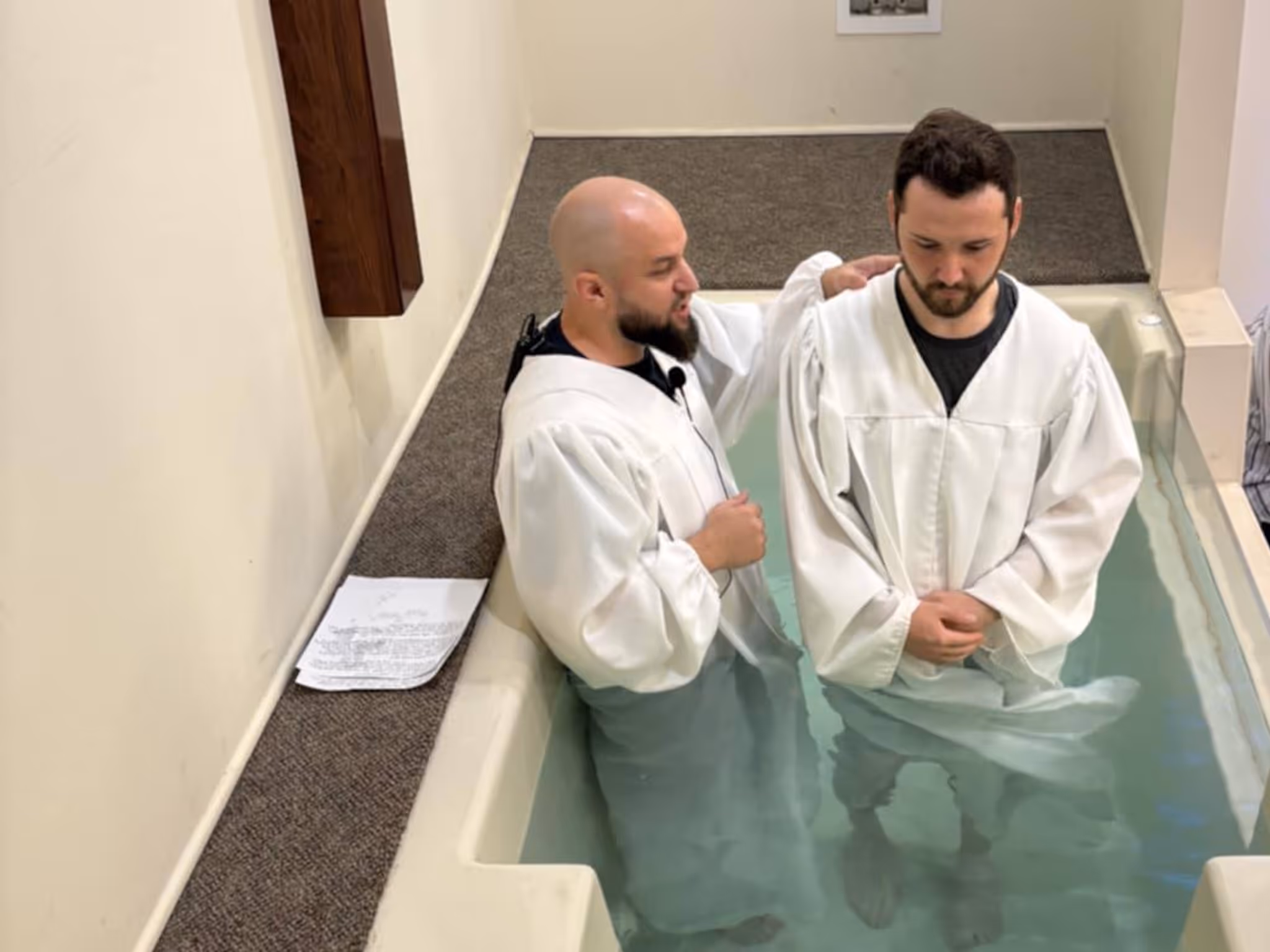 Two men in white robes stand in a baptismal pool, one with hand on the other's shoulder.
