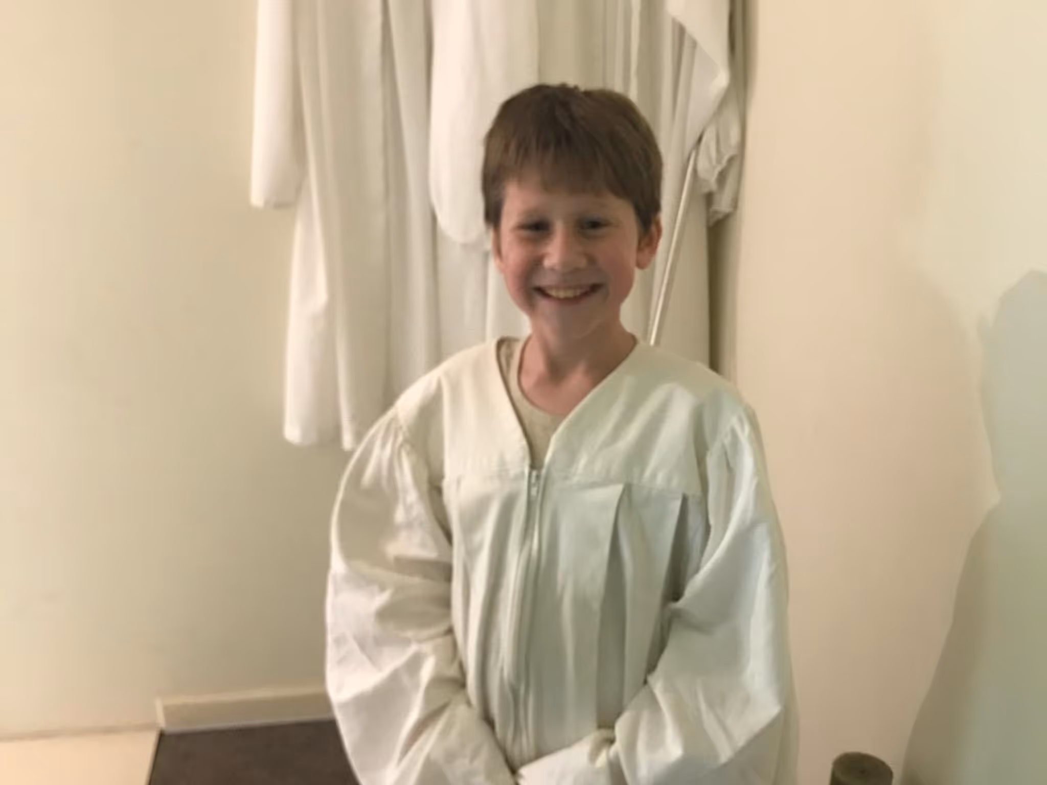 Smiling boy wearing a white baptism robe standing indoors in front of hanging robes.