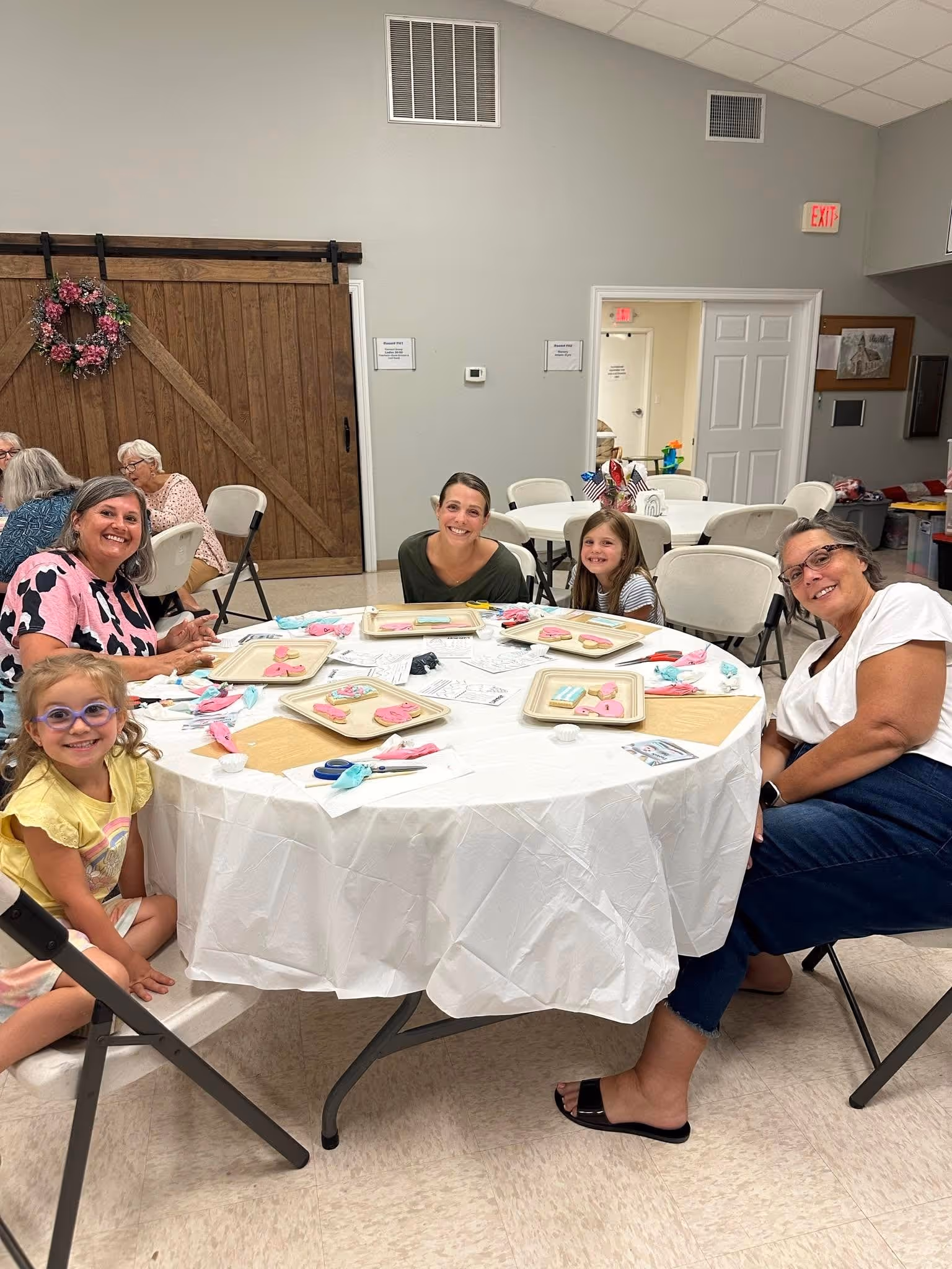 Smiling adults and children seated around a table with decorated cookies and icing bags inside a community room.