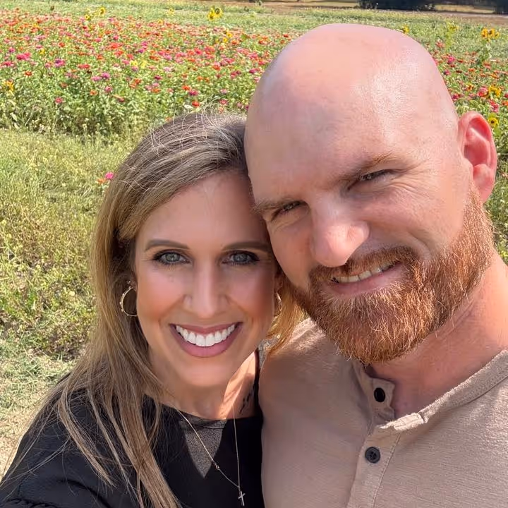 Smiling man with red beard and bald head and woman with blonde hair in a field of wildflowers.