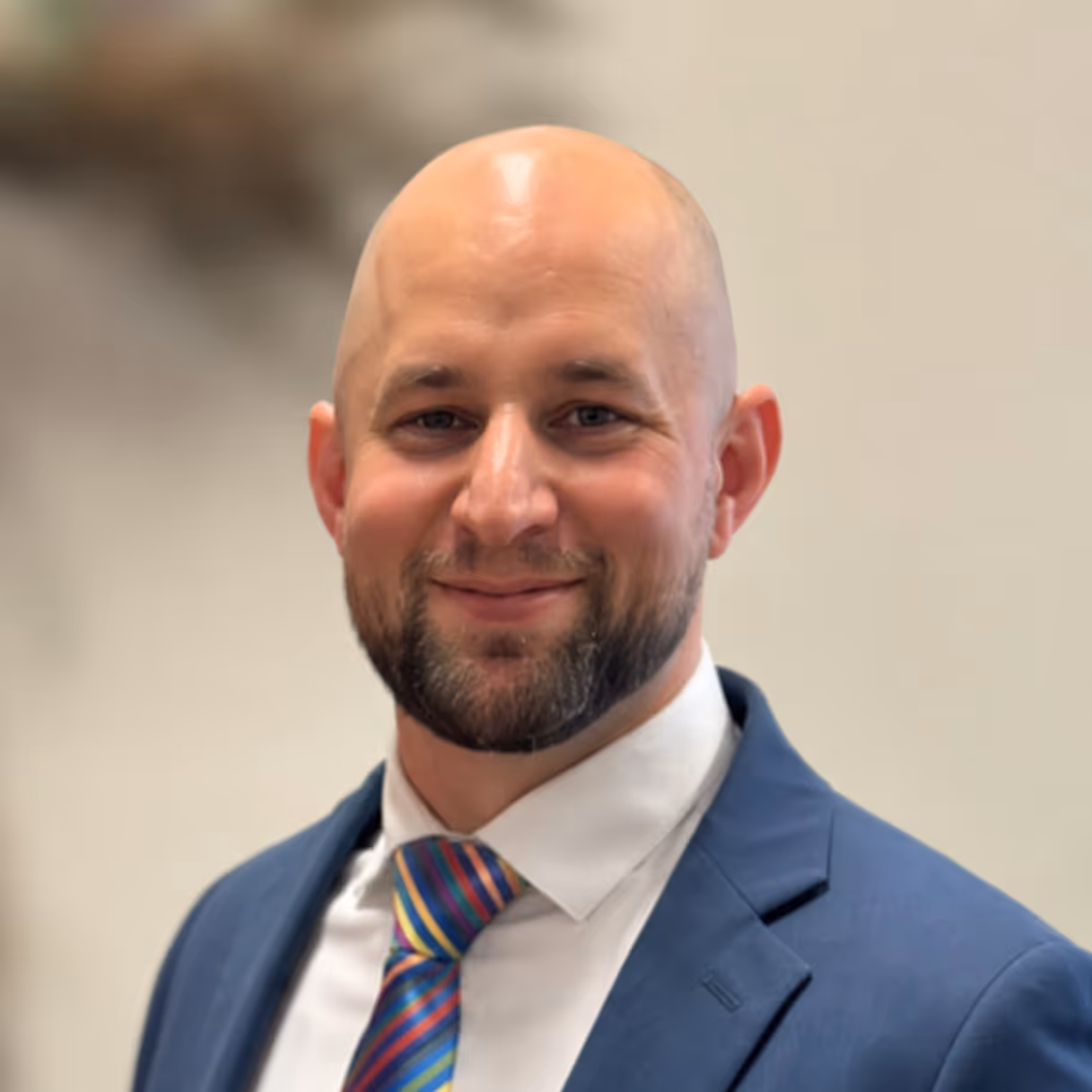 Smiling bald man with beard wearing a blue suit and colorful striped tie on a blurred background.