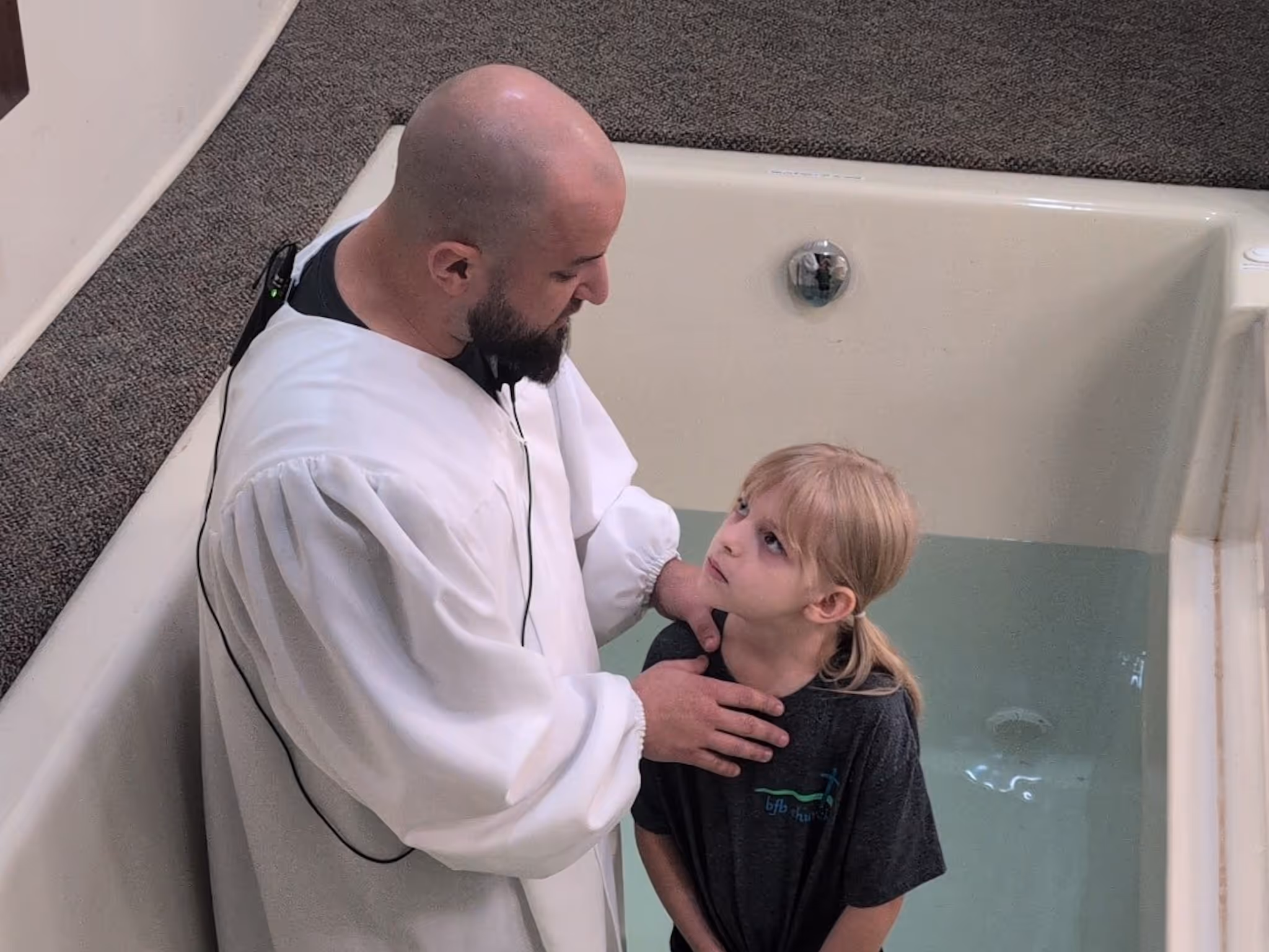 Man in white robe baptizing young girl standing in a baptismal pool filled with water.