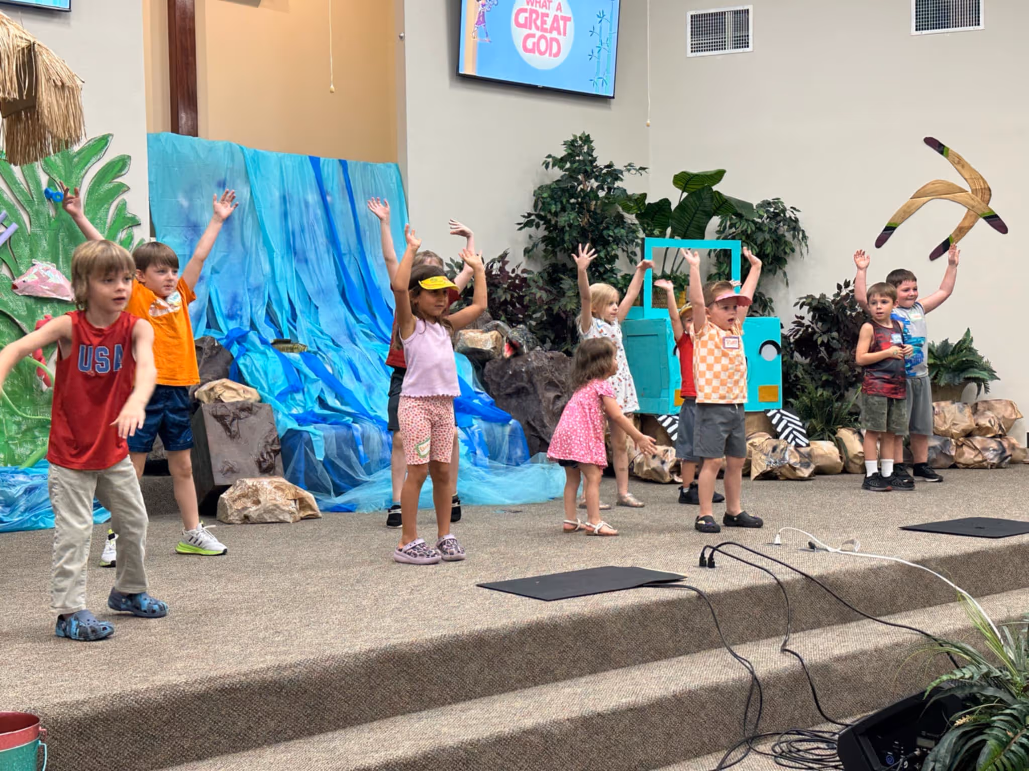Group of children on stage performing with arms raised, colorful backdrop, and props behind them.