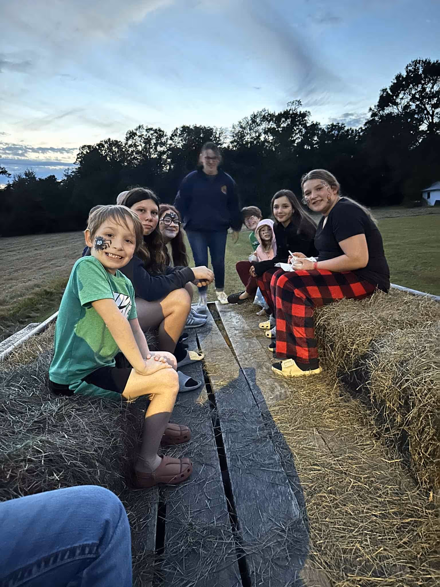 Group of children with face paint sitting on hay bales outdoors at dusk on a wooden platform.