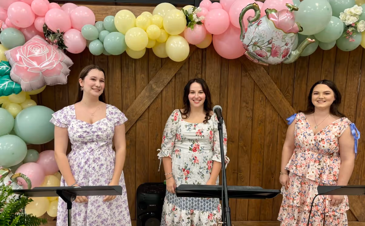 Three smiling women in floral dresses stand behind microphones with pastel balloon decorations overhead.
